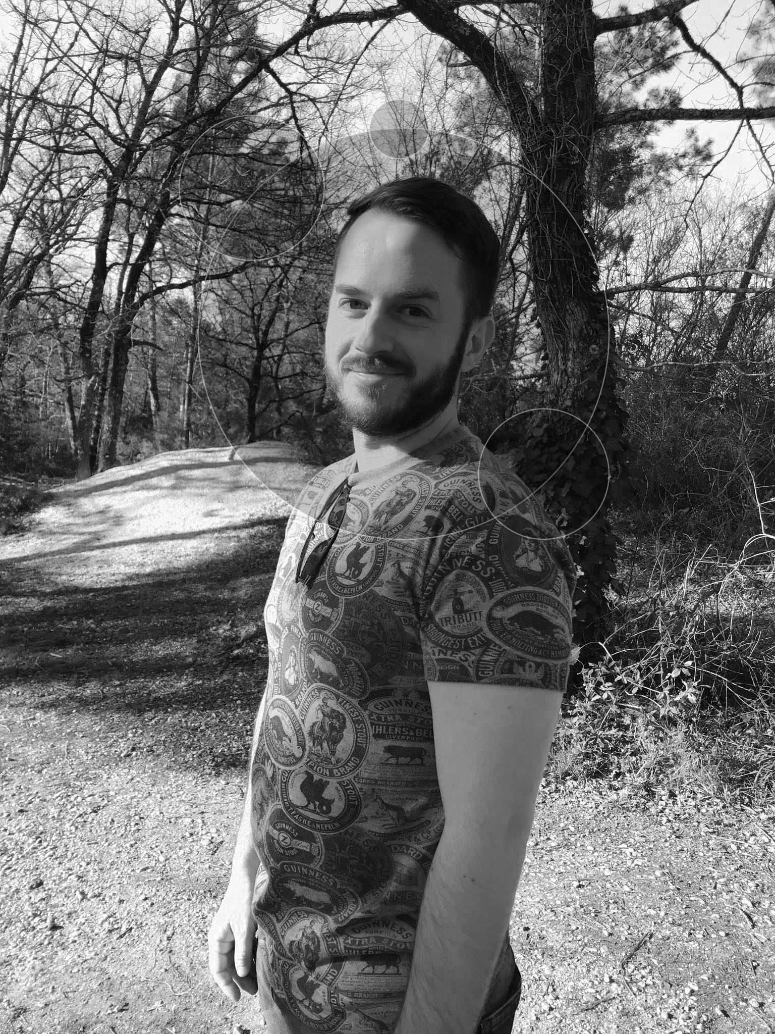 Fabien souriant à la caméra sur un chemin de forêt en noir et blanc.